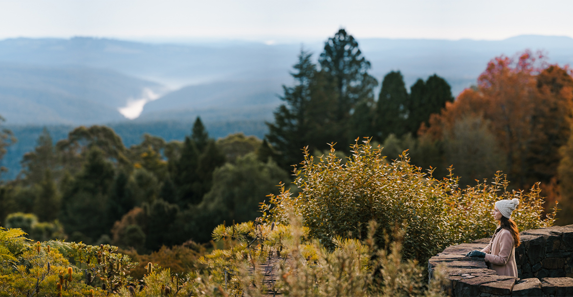 Woman in beanie and coat looks out over garden with mountain views and autumnal trees