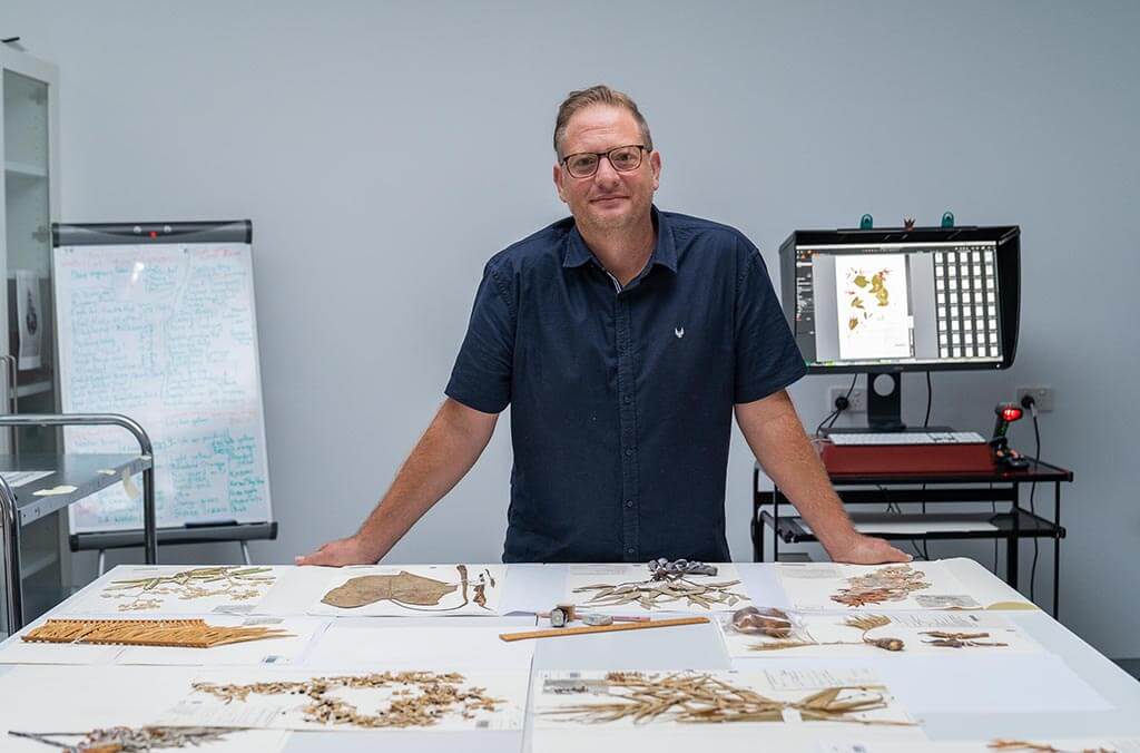 A scientist stands behind a table of herbarium specimens.